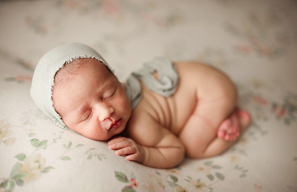 Sleeping newborn baby wearing a soft green bonnet posed on a floral blanket.
