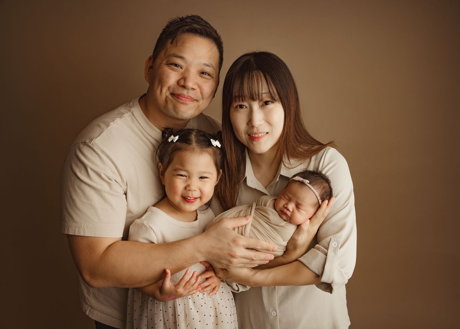 A happy Asian family of four poses for a professional portrait, featuring dad, mom, toddler daughter, and newborn baby.