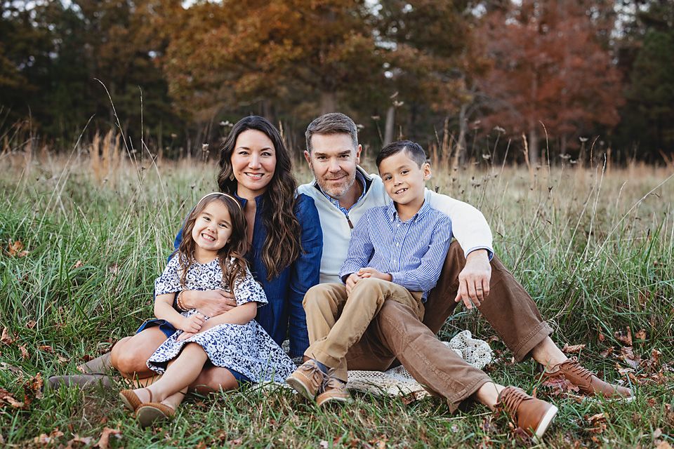 A family of four sits together smiling in a golden autumn field for a fall portrait.