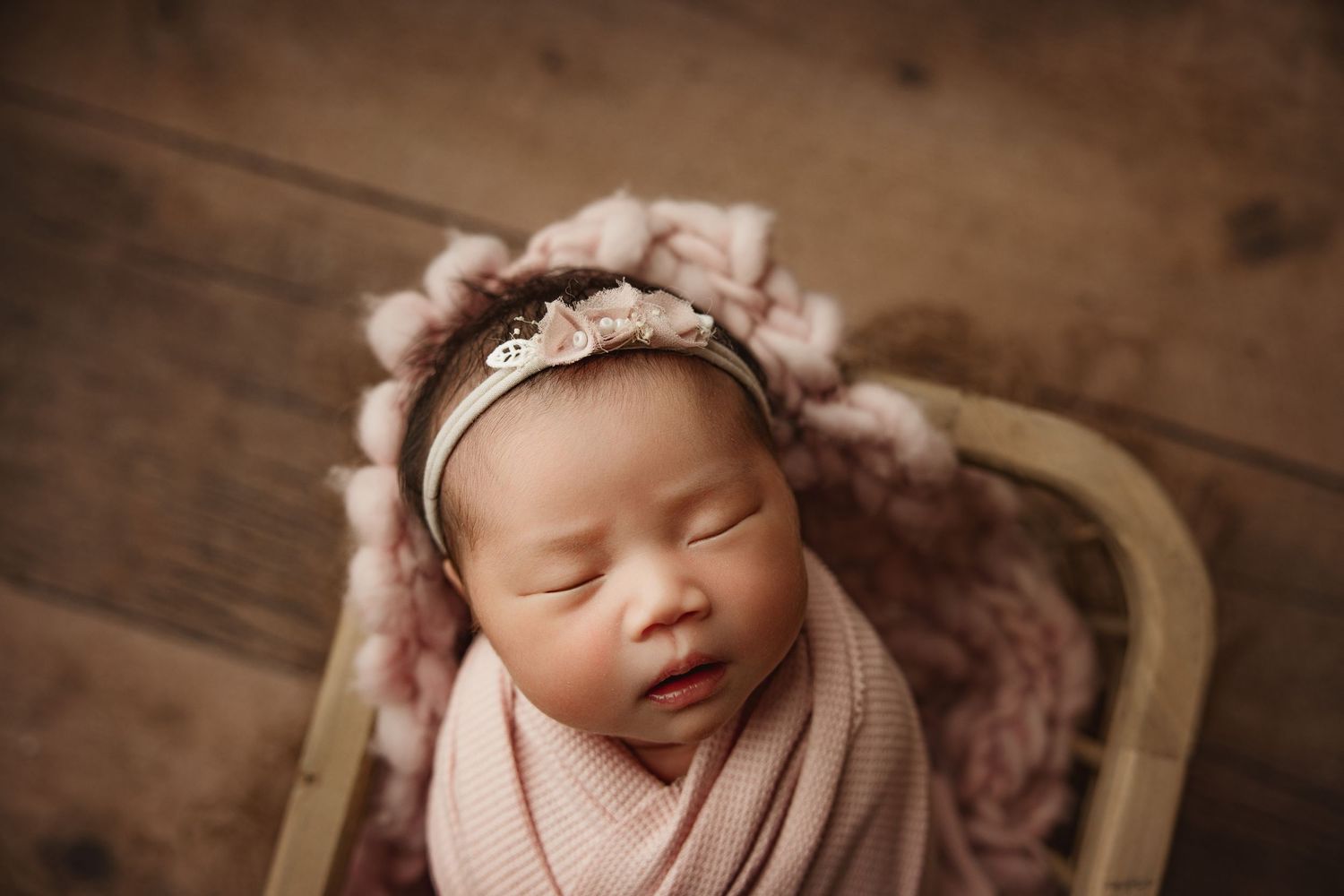 Sleeping newborn baby girl wrapped in pink swaddle with lace headband in a rustic wooden basket.