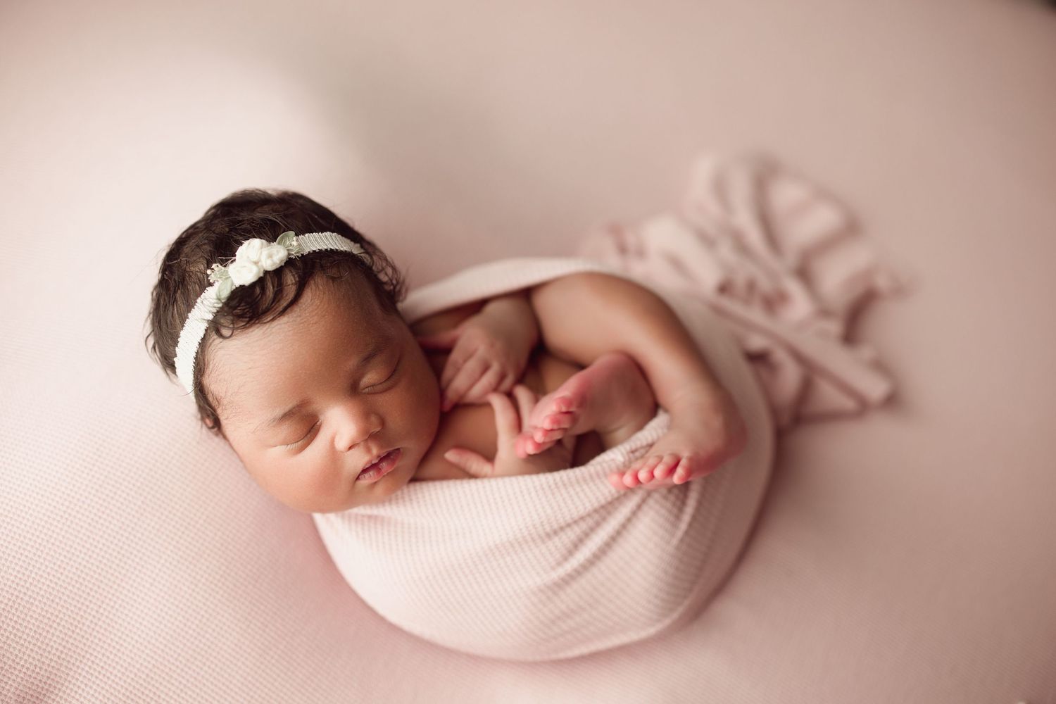 Sleeping newborn baby girl wrapped in pink swaddle with floral headband on pink background.