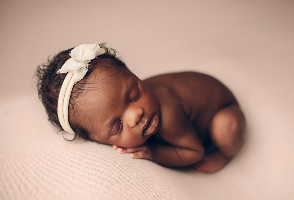 Adorable smiling newborn baby with white bow headband lying on soft pink blanket.