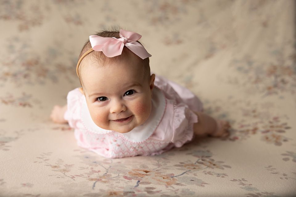 Smiling baby girl in pink dress with bow headband during tummy time portrait session.