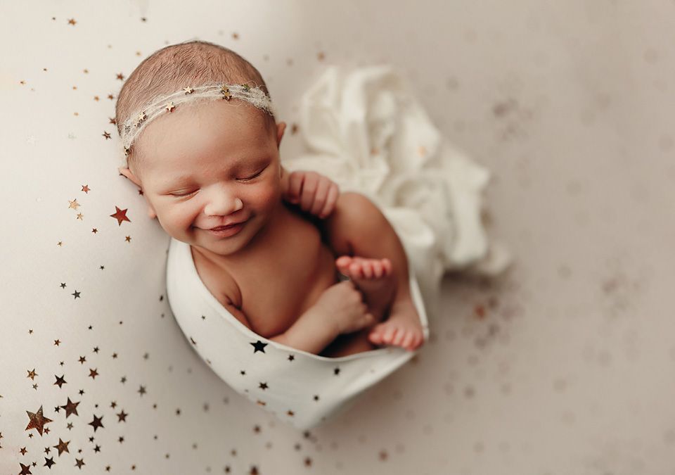 Smiling newborn baby wrapped in white cloth with star confetti on a soft background. Richmond Photography
