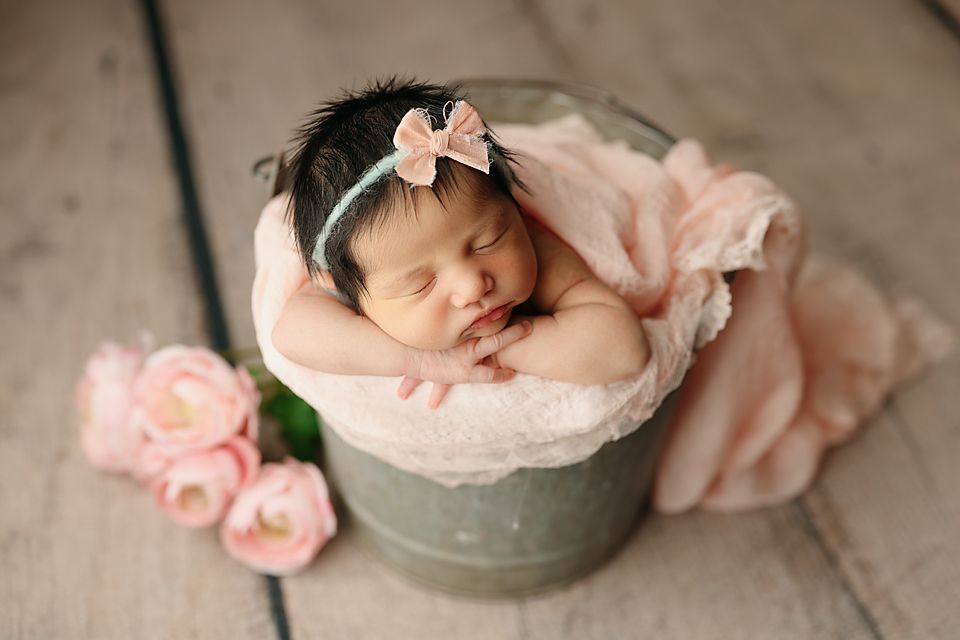 Sleeping newborn baby girl in a metal bucket with pink roses and a bow headband on a wooden floor Richmond newborn photographer