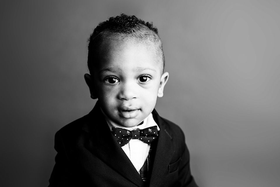 Adorable toddler boy in a black suit and polka dot bow tie poses for a formal black and white portrait.