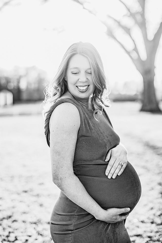 Black and white photo of a smiling pregnant woman holding her belly outdoors among fallen leaves. New Kent maternity photographer