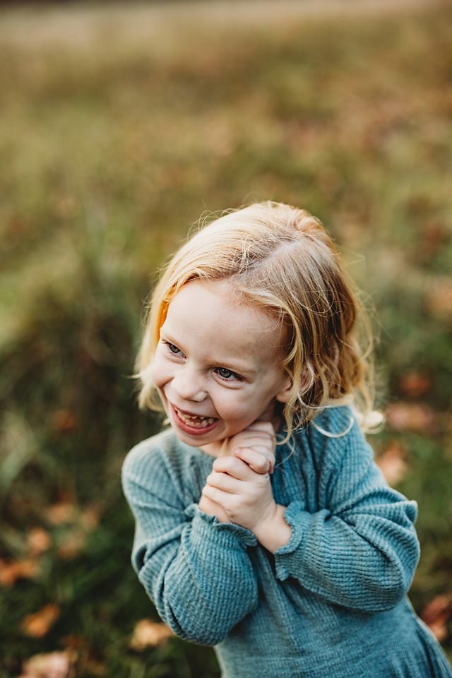 A cute blonde toddler girl in a teal sweater makes a playful face outdoors in fall.