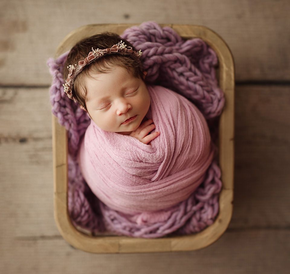 Newborn baby girl wrapped in pink, sleeping in a rustic wooden box with a pretty floral headband.