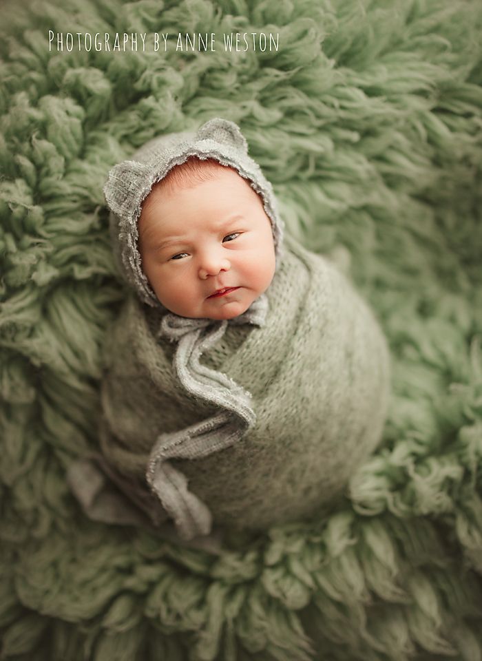 Newborn baby wrapped in a green knit swaddle with bear ear bonnet on a fluffy green backdrop.