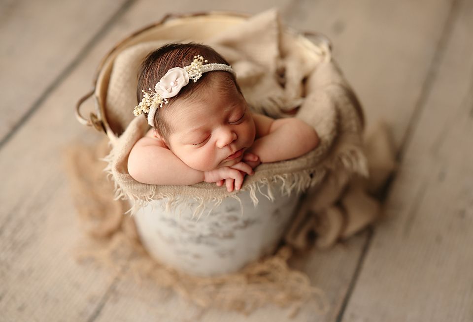 Sleeping newborn baby girl in a rustic bucket with a floral headband during a studio photo session.
