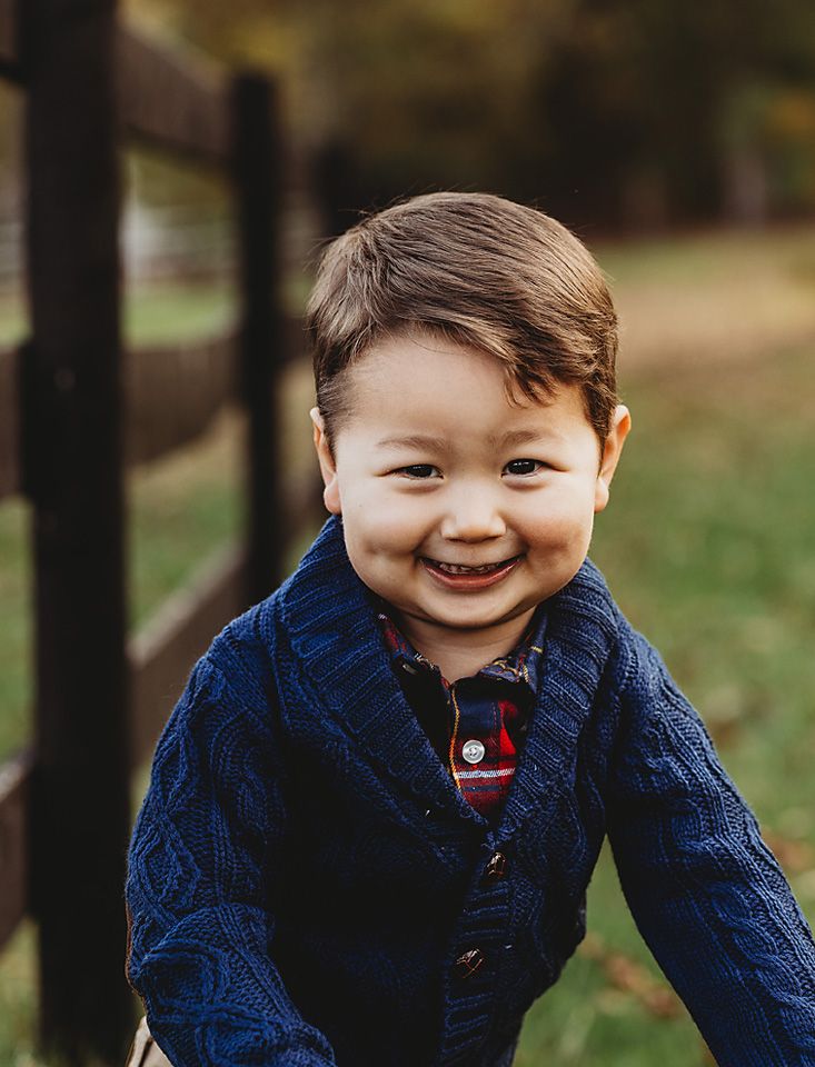An adorable toddler boy in a navy blue sweater smiles brightly outdoors in fall.
