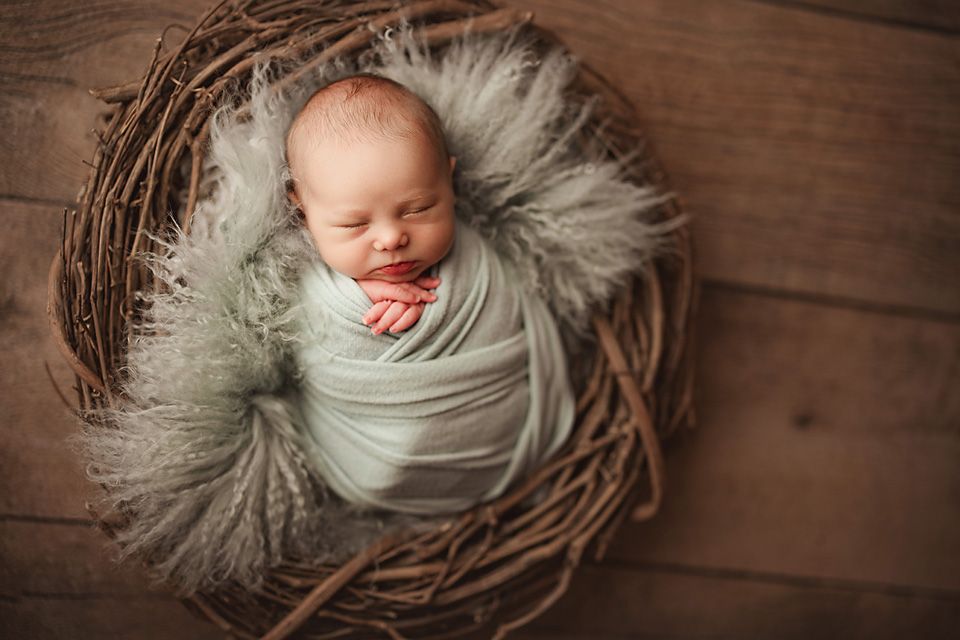 Sleeping newborn baby wrapped in mint swaddle nestled in a rustic twig nest with fluffy fur.