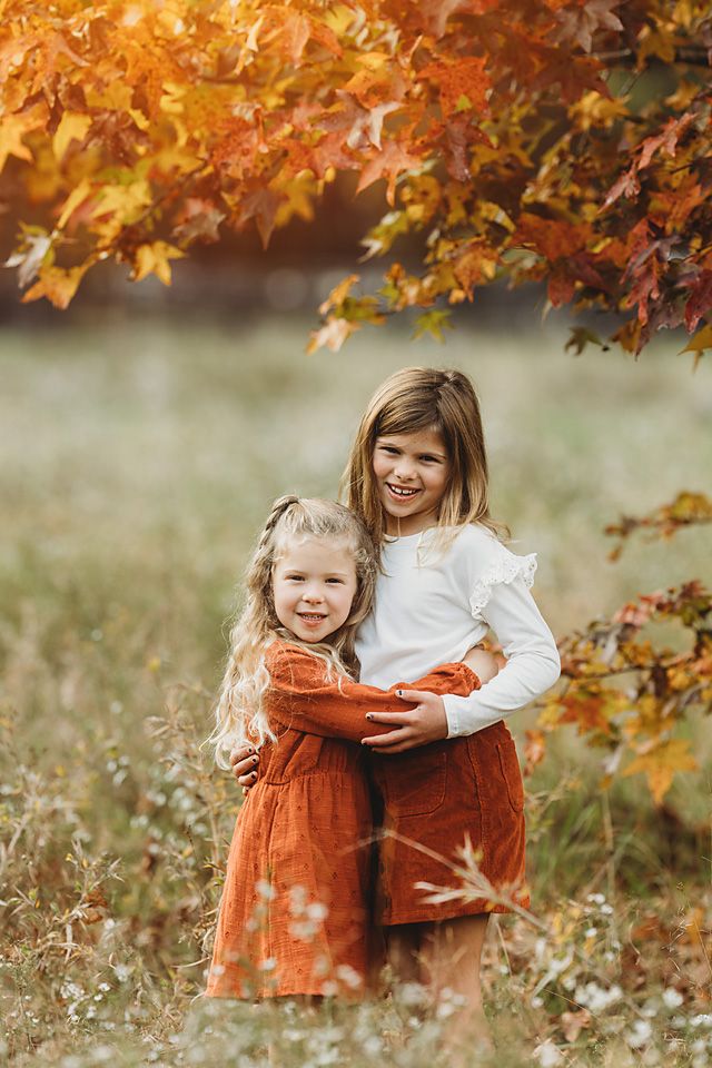 Two young sisters in rust and white dresses embrace under vibrant orange autumn maple tree leaves Richmond VA family photography. Midlothian