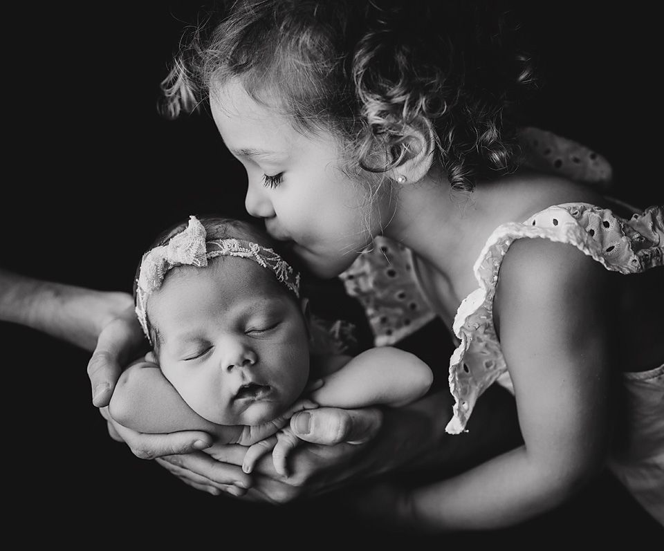 Black and white photo of a toddler girl kissing a sleeping newborn baby cradled in adult hands.