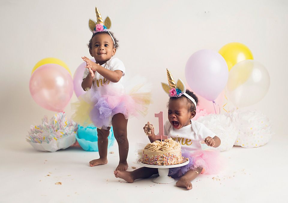 Two toddler girls in unicorn tutus celebrating first birthday with cake and colorful balloons.