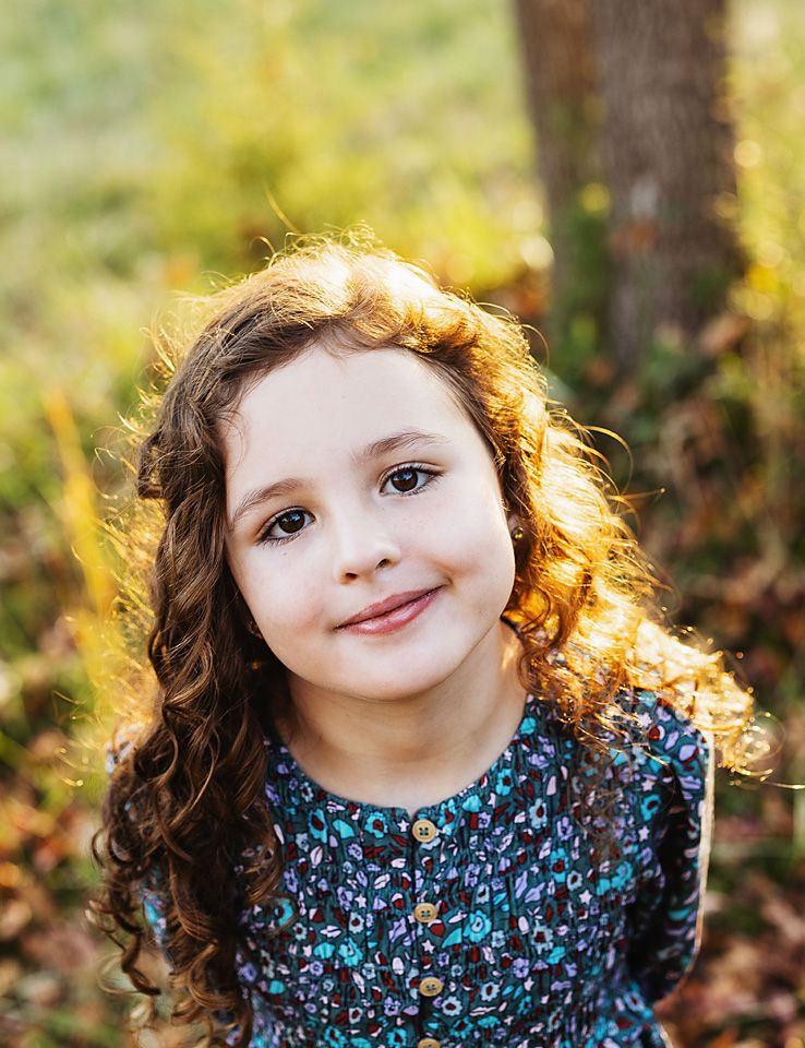 Young girl with curly brown hair smiles softly in a floral dress during golden hour outdoor portrait.