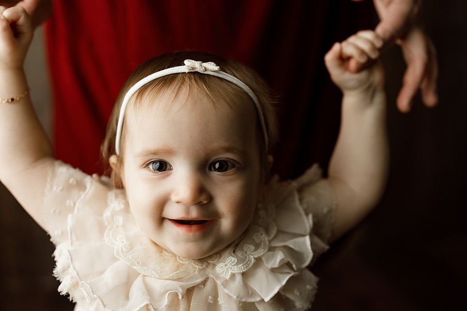 Smiling baby girl in white lace dress with bow headband reaching up during portrait session.