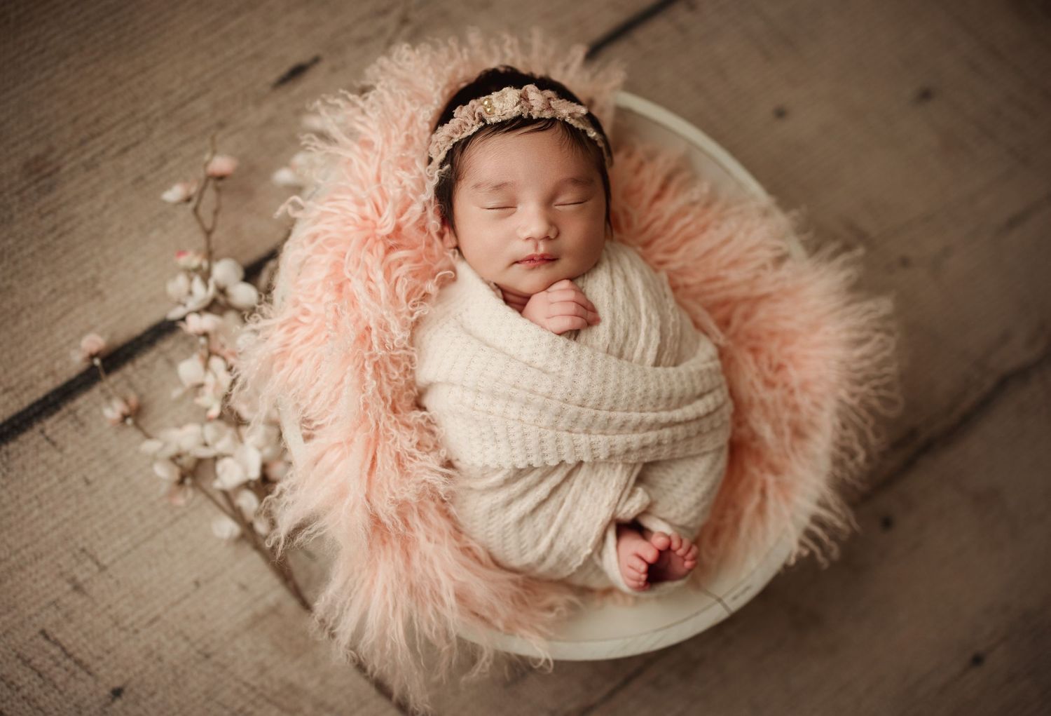 Newborn baby girl swaddled in cream wrap, sleeping in a bowl with pink fluffy fur, wearing a floral headband.