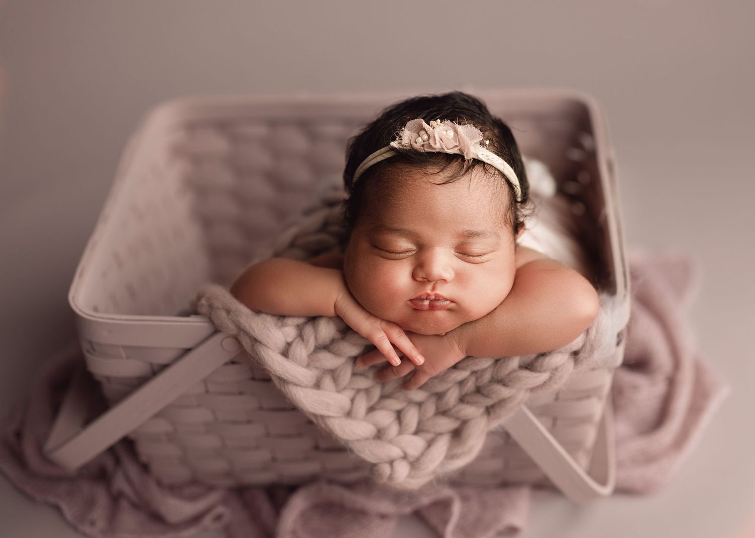 Newborn baby girl sleeping peacefully in a gray basket with chunky knit blanket and floral headband.