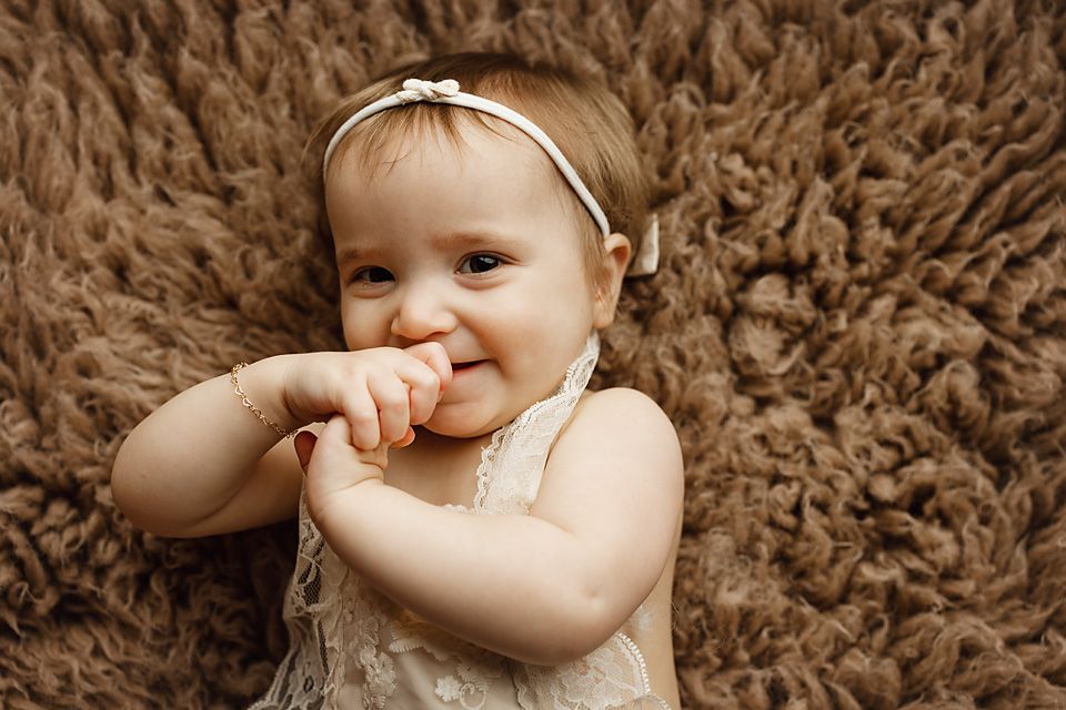 Sweet baby girl in white dress lying on fluffy brown rug chewing fingers during photo session.