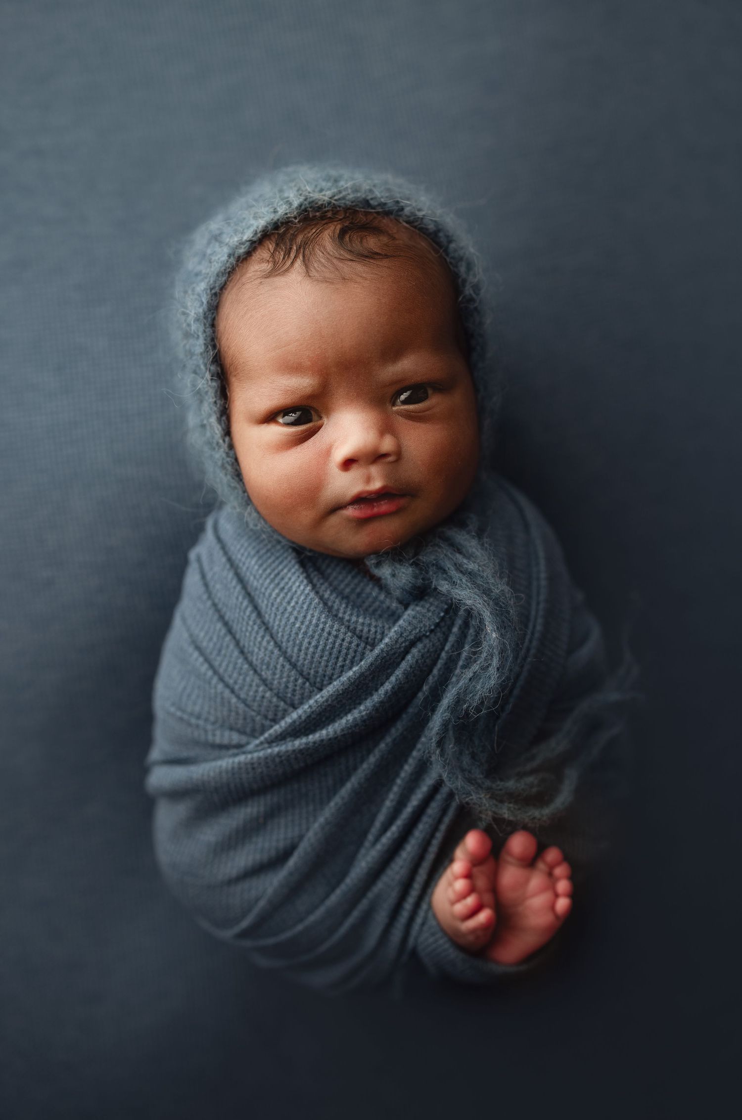Newborn baby swaddled in a teal knit wrap with a matching bonnet on a dark blue background.