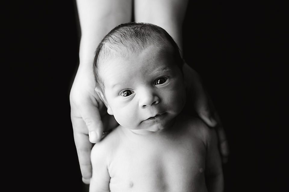 Black and white newborn portrait with adult hands cradling alert baby against dark background.
