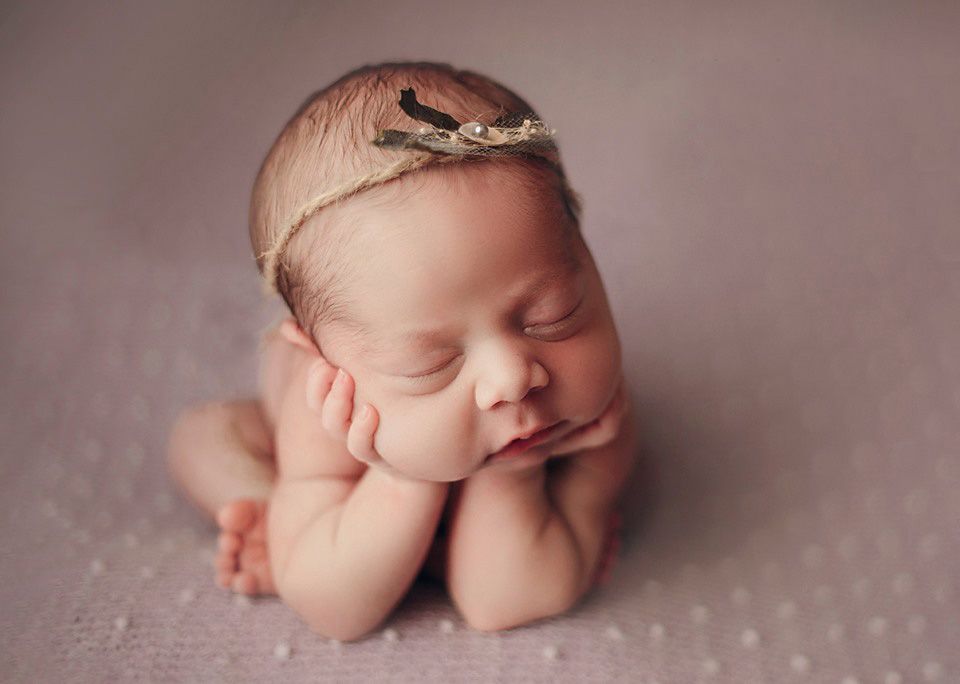 Peaceful sleeping newborn baby posed with hands under chin wearing delicate gold tiara headband.