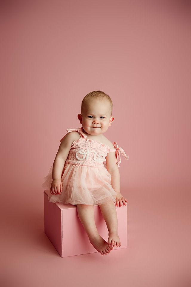 Baby girl in pink tutu dress sitting on pink cube for first birthday photo session.