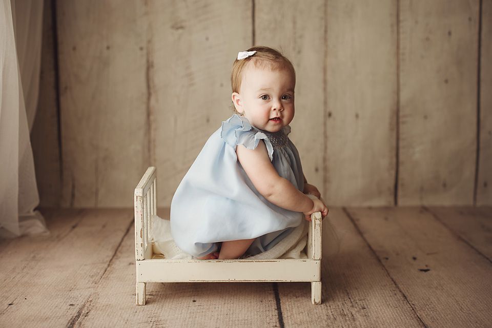 Baby girl in a blue dress sits on a miniature wooden bed on rustic hardwood floors.