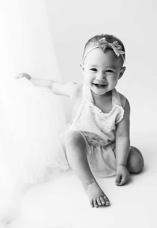 Happy baby girl in white tutu dress with sparkle bow headband laughing in black and white photo.