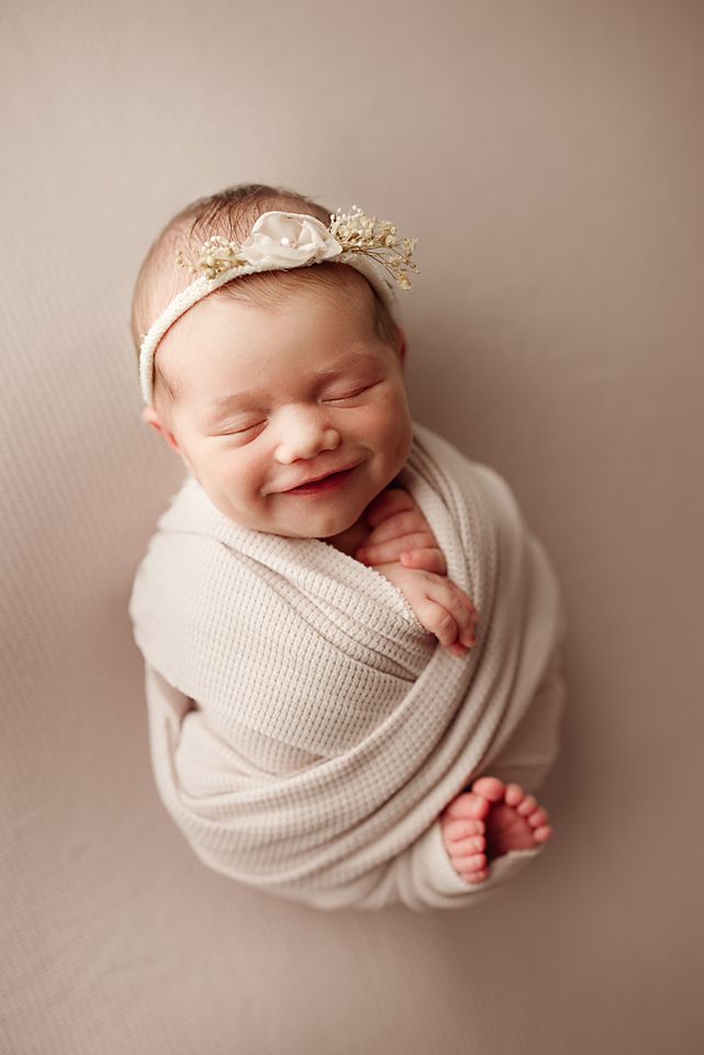 Smiling newborn baby girl swaddled in cream wrap wearing floral headband, posed on beige backdrop.