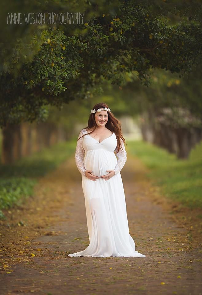 Pregnant woman in white lace gown and floral crown stands on tree-lined path in a maternity photo by Anne Weston Photography.