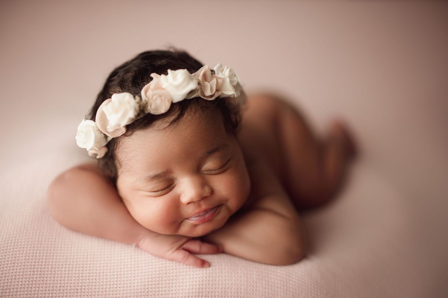 Smiling newborn baby girl wearing a white floral crown, peacefully resting on a soft pink blanket.
