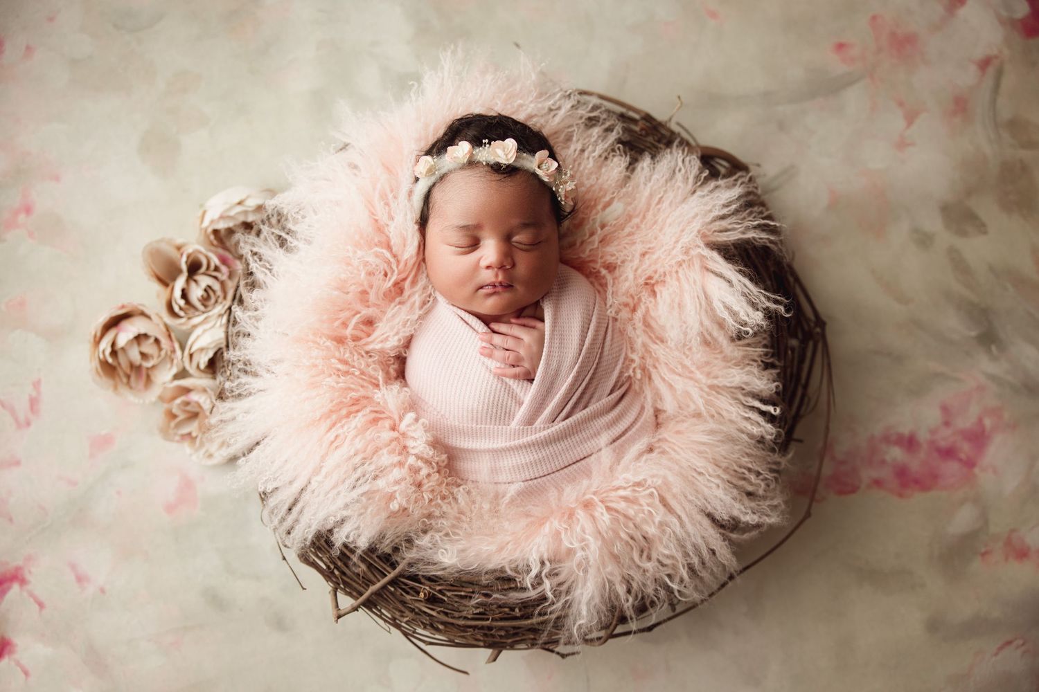 Newborn baby girl wrapped in pink sleeping in a rustic basket with fluffy pink feathers and a floral headband.