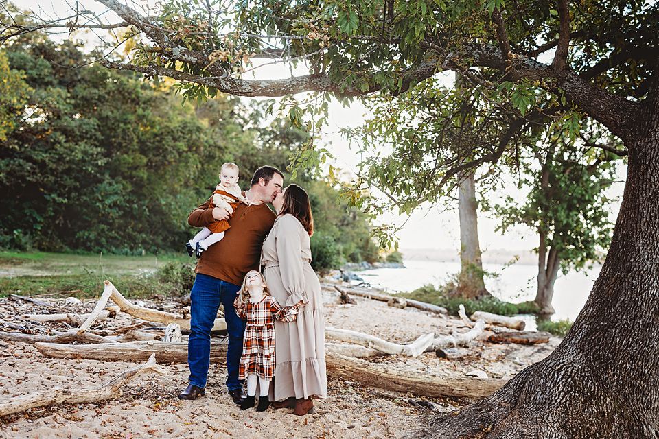 Family of four sharing a kiss near driftwood beach under large tree.