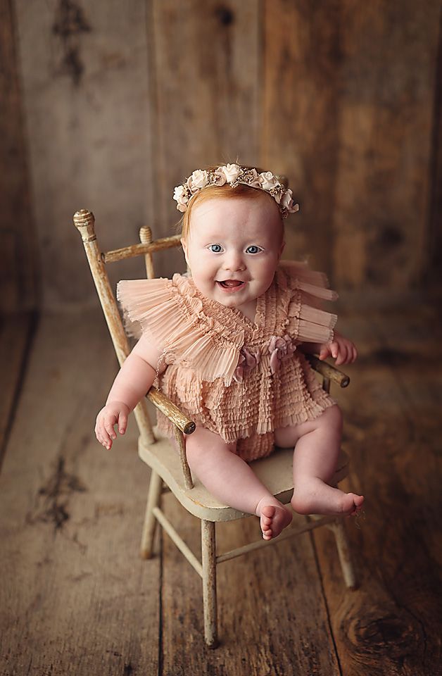 Happy baby girl in peach ruffle romper with floral crown sitting in vintage chair portrait.