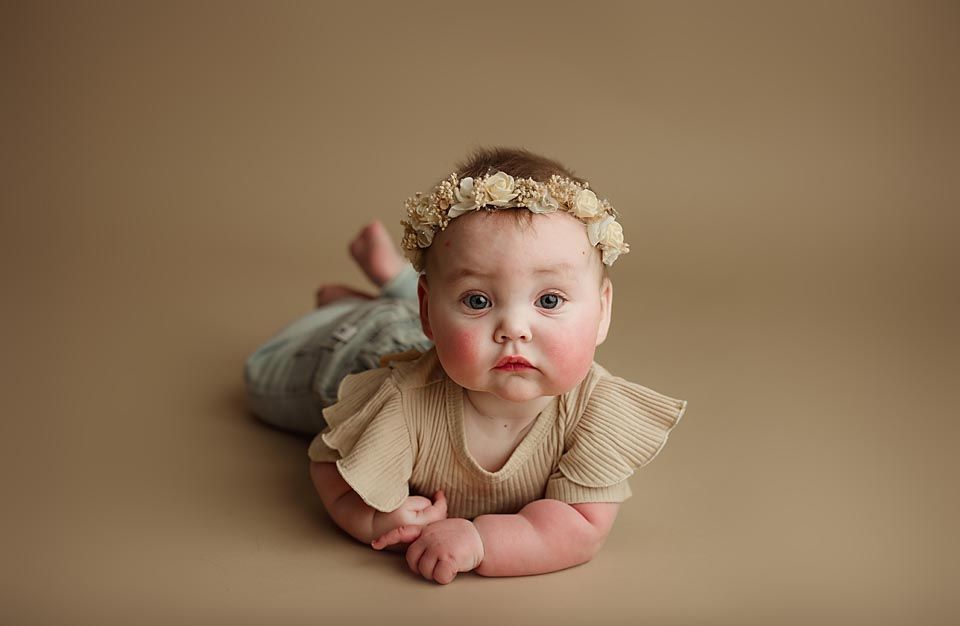 Cute baby girl in beige ruffled outfit with floral headband doing tummy time on a tan background.