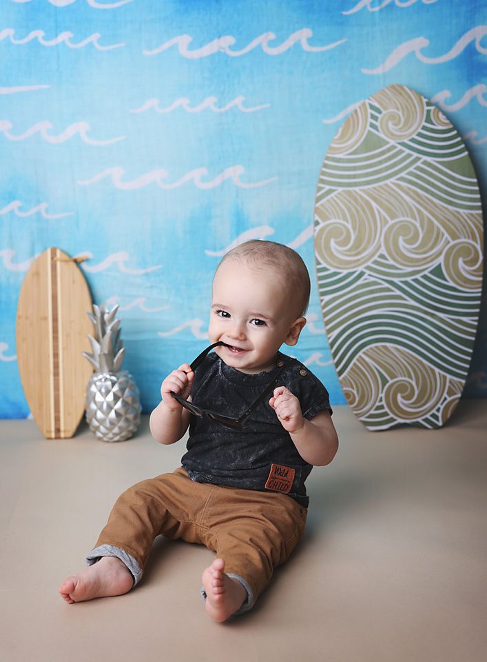 Smiling baby boy sitting with surfboards and tropical decor on a beach-themed backdrop.