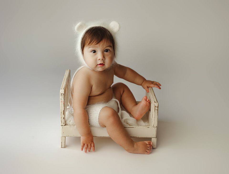 Cute baby in white bear ear bonnet sitting on miniature vintage white wooden bed against gray backdrop.