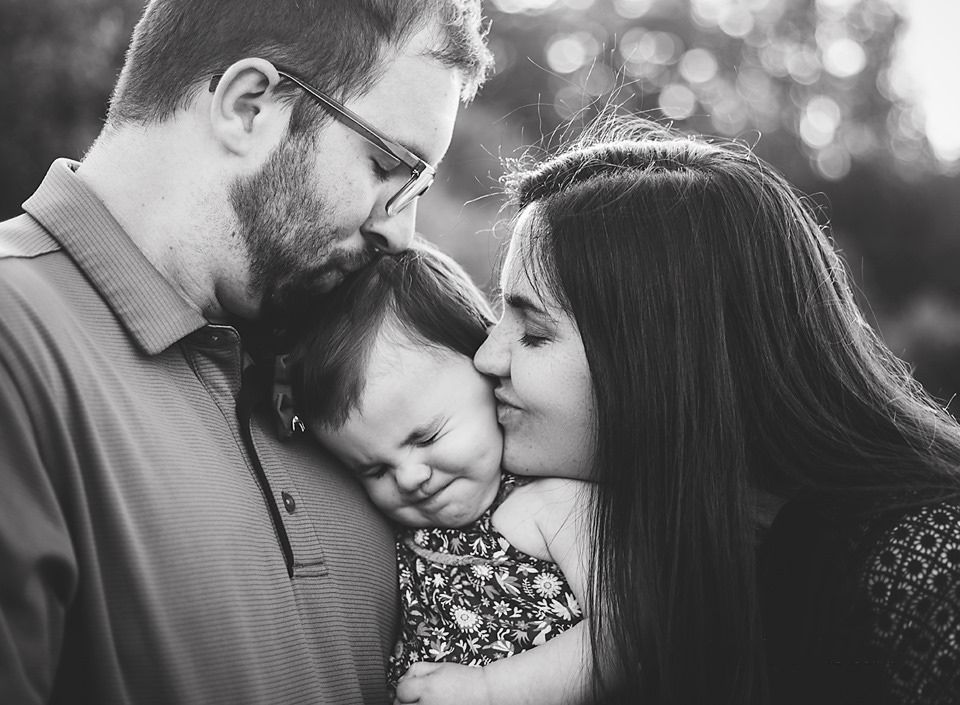 Black and white photo of loving parents kissing their baby on the cheeks outdoors.