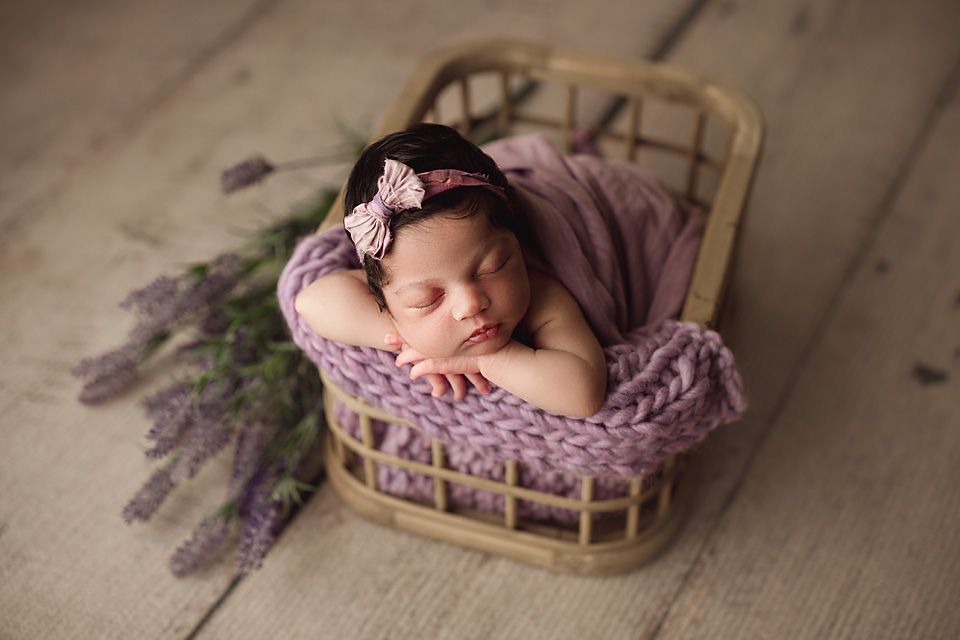 Sleeping newborn baby girl in purple basket with lavender flowers and bow headband.