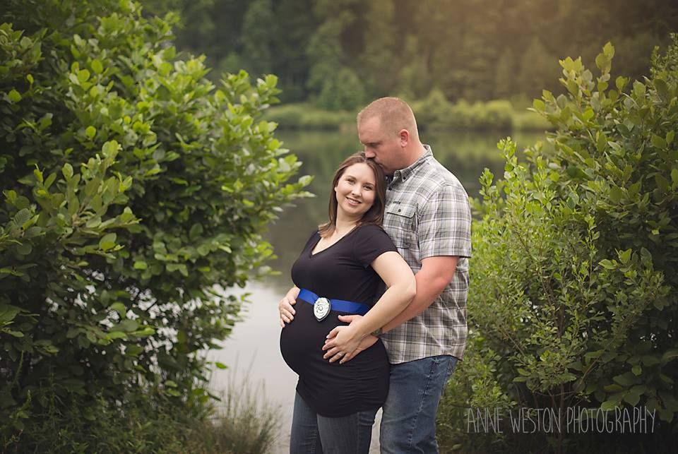 Couple poses outdoors during maternity photo shoot, woman in black top showing baby bump, surrounded by lush green trees. Richmond