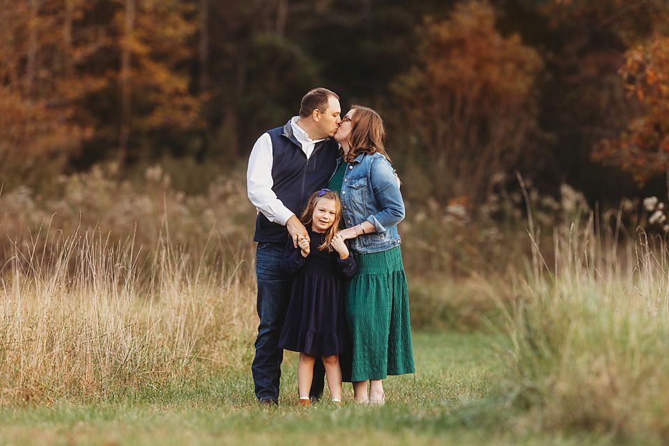 Couple sharing a kiss while their young daughter stands between them in fall field.