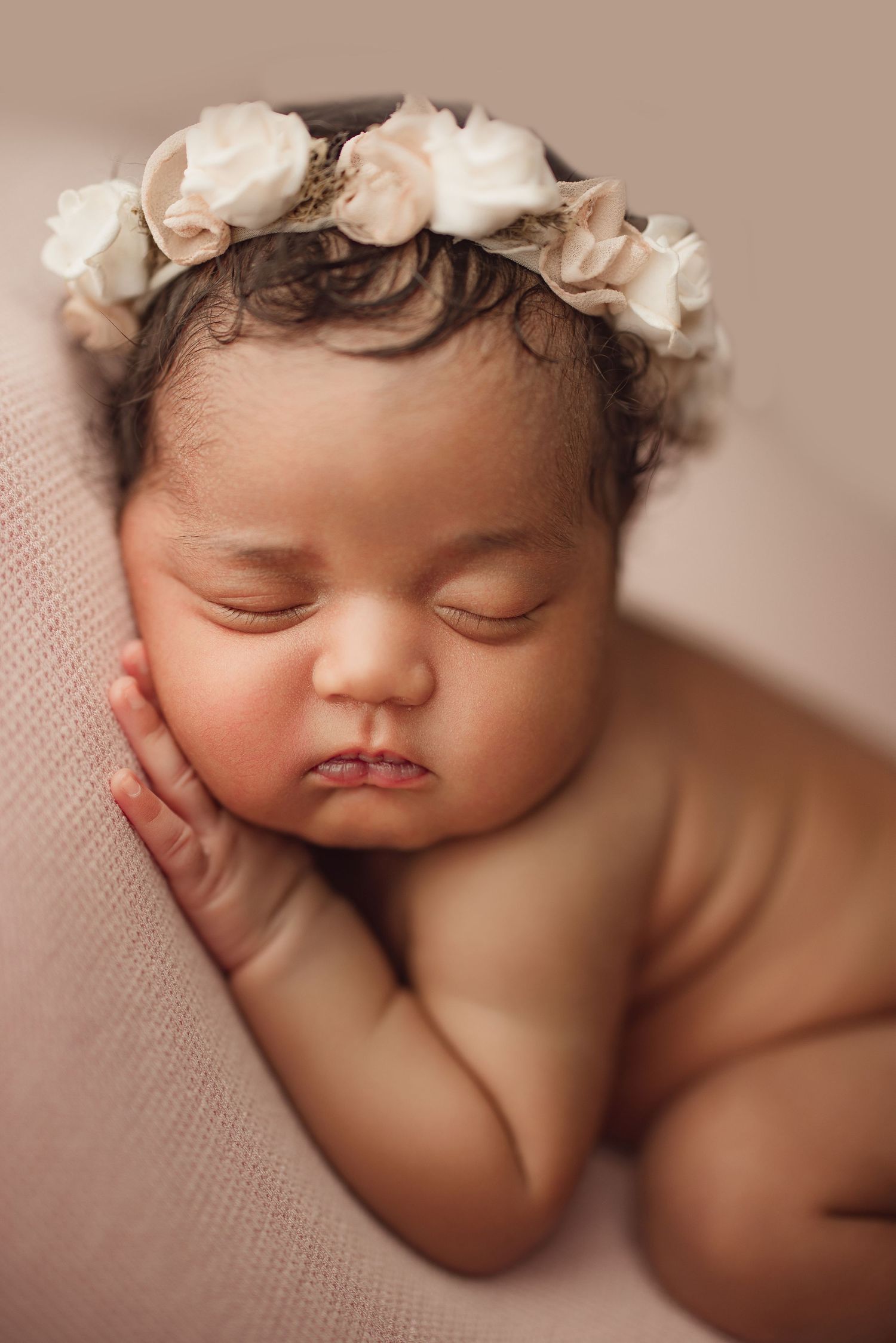 Sleeping newborn baby girl wearing a delicate white floral crown, resting peacefully on a soft pink blanket.