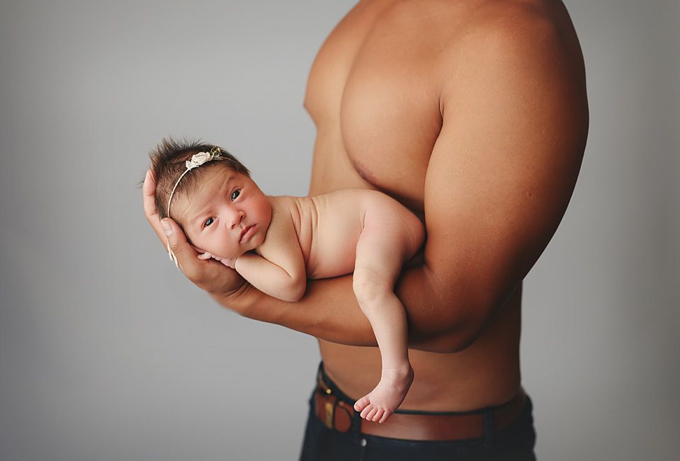 Shirtless father tenderly holding a sleeping newborn baby girl with a headband against a gray background Richmond newborn photographer