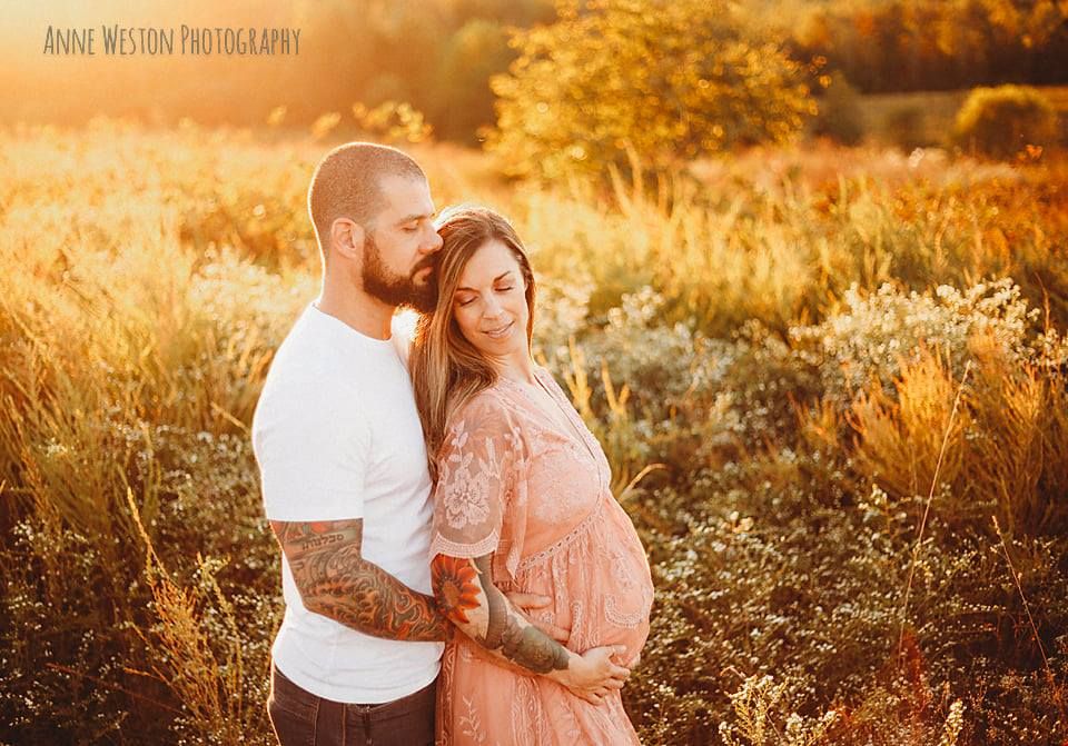 Couple sharing a tender maternity moment in a golden sunlit field, woman in pink lace dress cradling baby bump. Goochland, VA