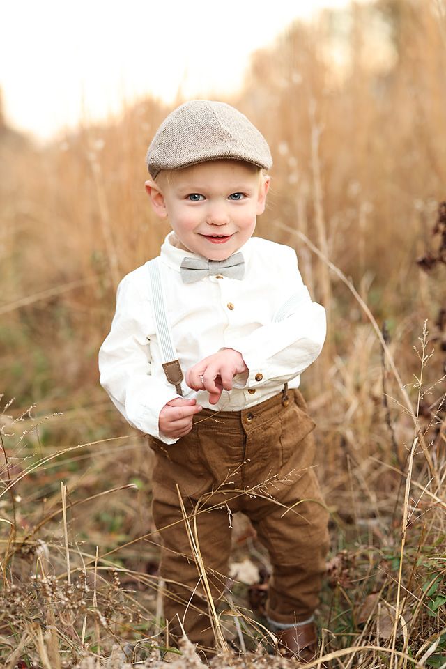 Adorable toddler boy in vintage suspenders, bow tie, and flat cap posing in a golden field.