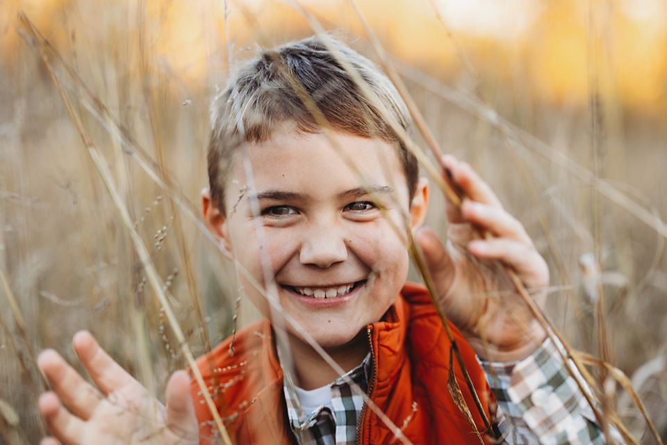 Smiling boy in an orange vest laughs joyfully while standing in tall golden grass during fall portraits. mechanvicsville photographer