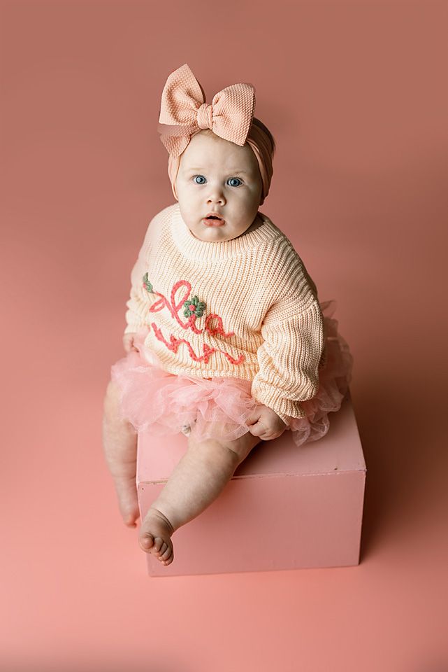 Baby girl in cream knit sweater and pink tutu sitting on a pink box against a pink backdrop.